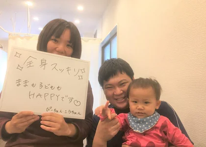 「全身スッキリ ままも子どももHAPPYです」と書かれたボードを持つ女性と、お子さんを抱いた院長のツーショット写真