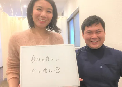 「身体の疲れは心の疲れ」と書かれたボードを持つ女性と、院長のツーショット写真